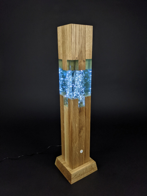 Lampa z dubového dřeva a světle modré epoxidové pryskyřice. V pryskyřici je zalitý světelný řetěz a lampa je rozvícená. Foceno na černím pozadí.